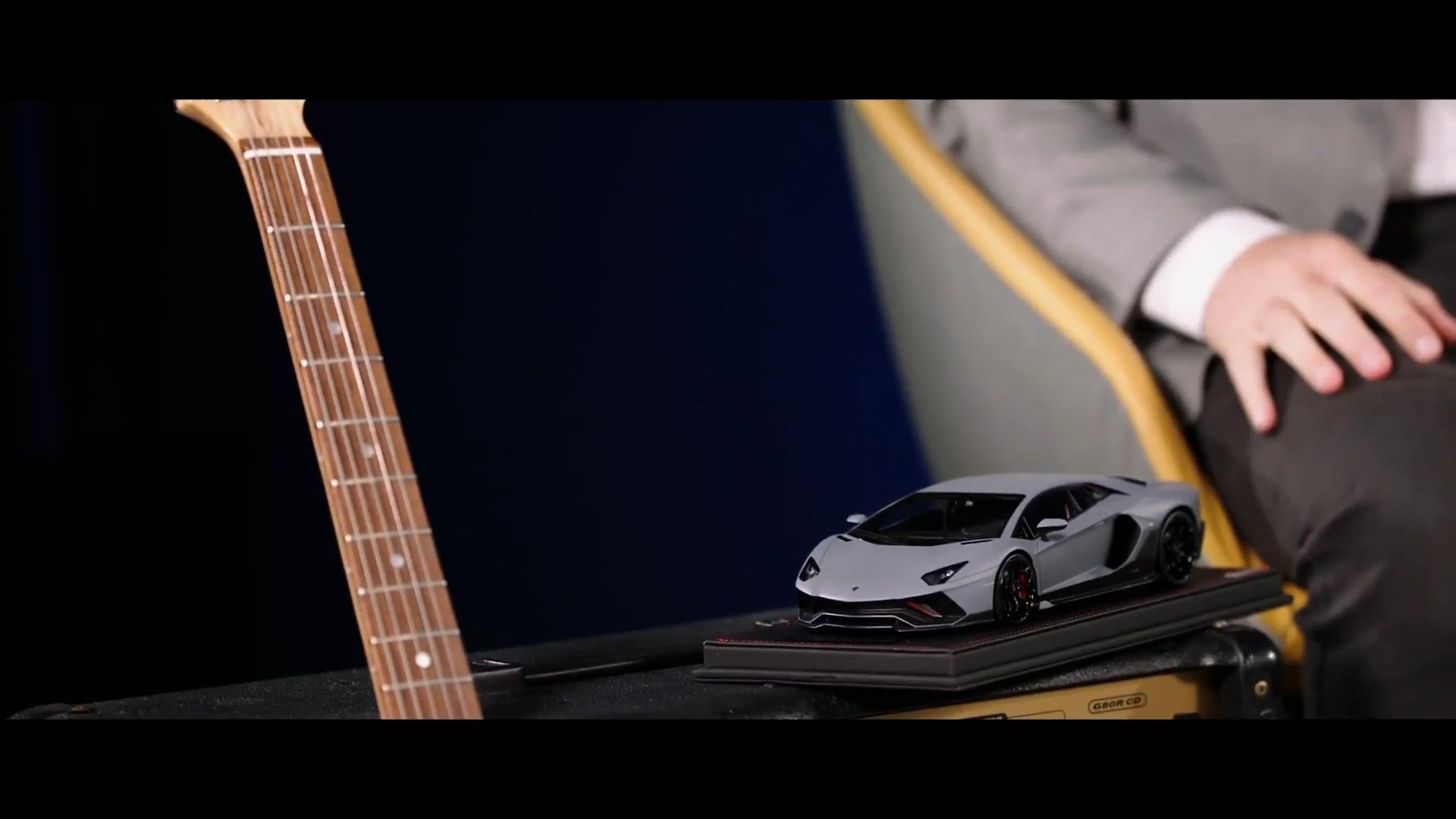 A scale model of a grey Lamborghini Aventador next to the neck of an electric guitar.