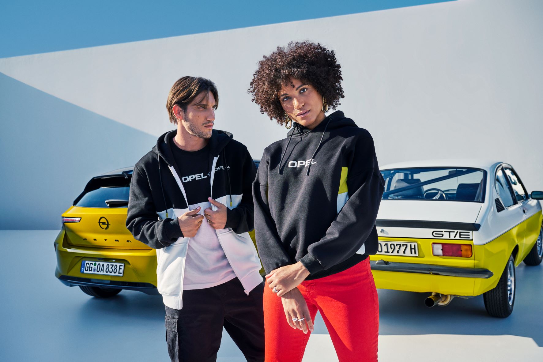 Two models wearing Opel clothing in front of a new Opel Astra and classic Opel Astra GTE both in yellow colours.