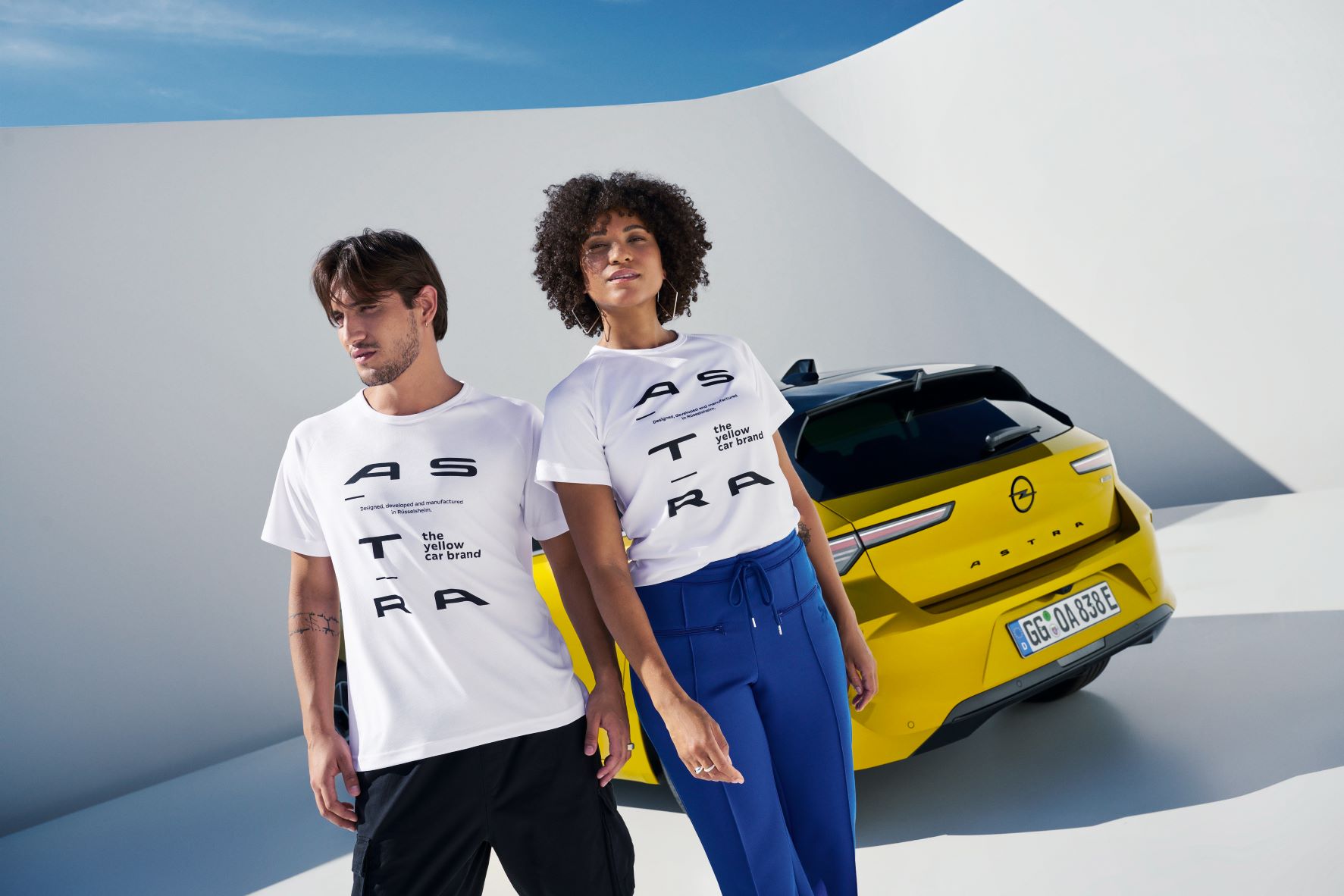 Two models wearing white Opel Astra t-shirts pictures in front of a Kult Yellow Opel Astra hatchback.