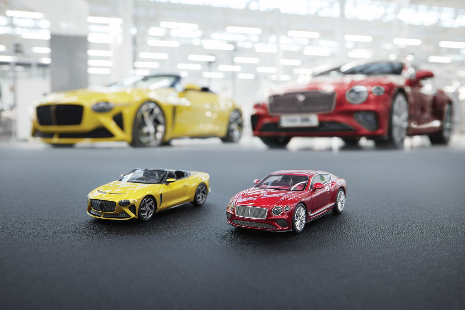 A yellow Bentley Bacalar model car and a red Bentley Continental GT Speed model car in front of the life sized cars.
