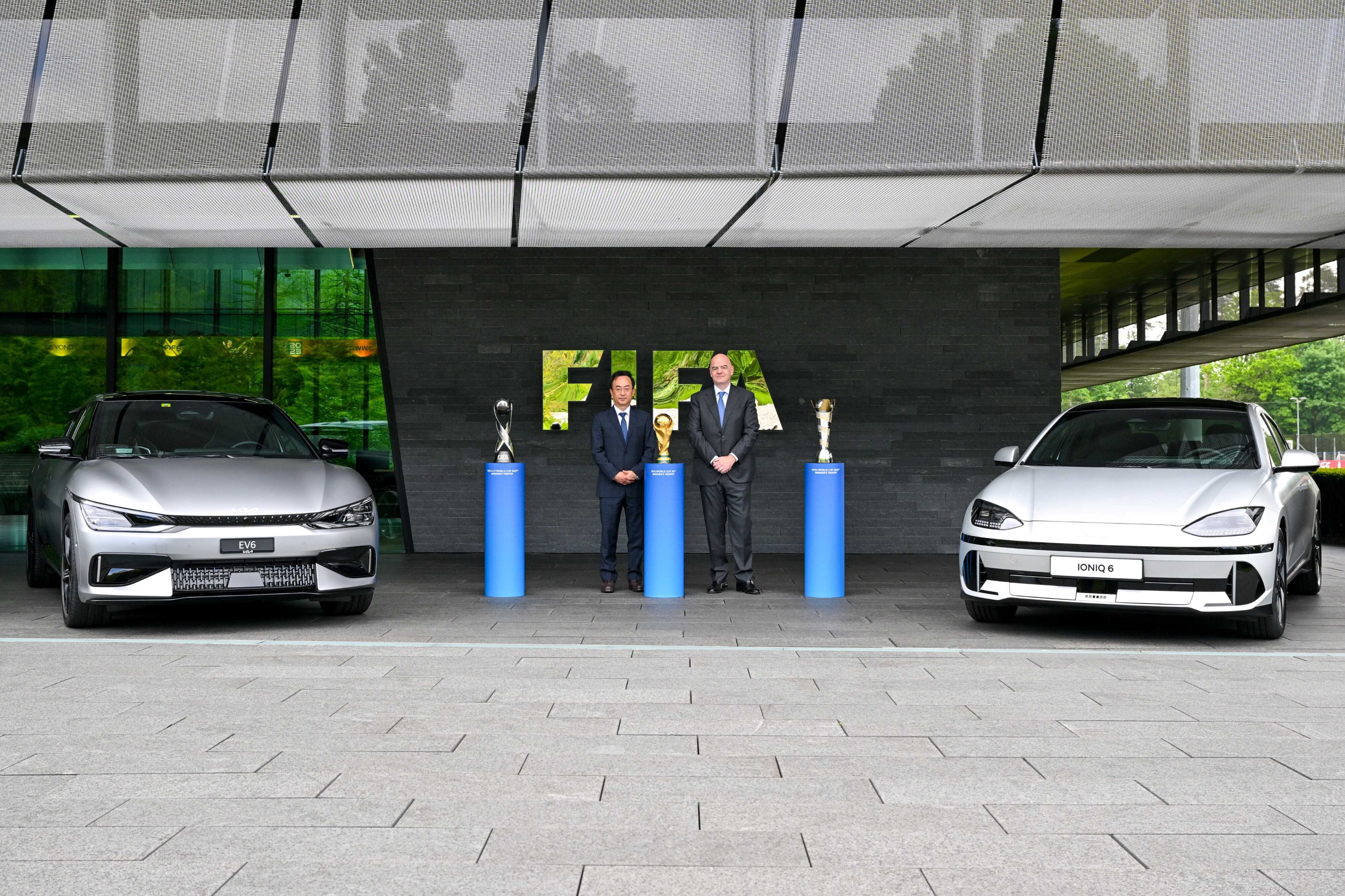 Representatives from Hyundai Motor Group and FIFA pose with the trophies for the football World Cup alongside a Hyundai IONIQ 6 in white and a KIA EV6 GT in grey.