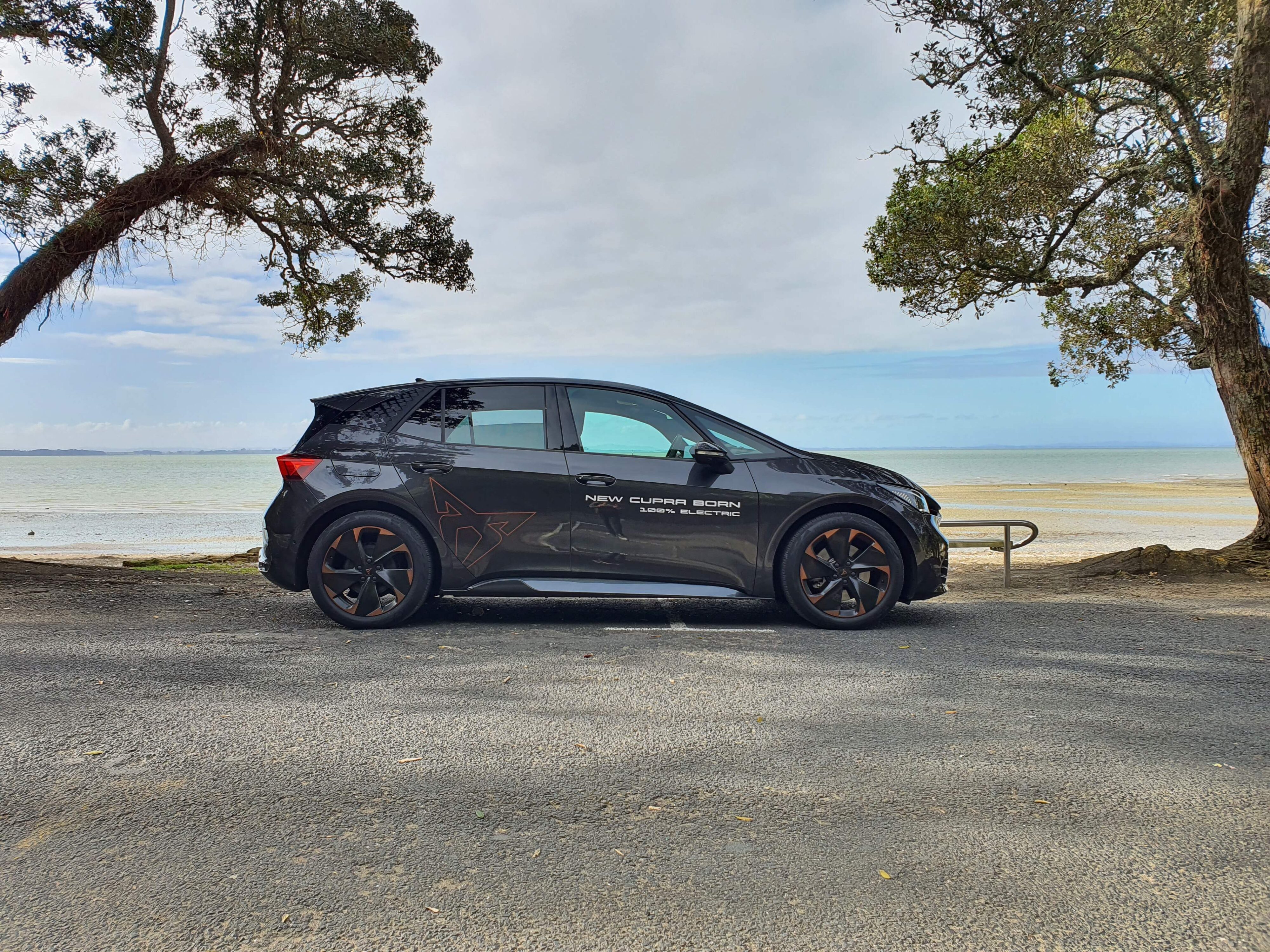 Side view of a 2023 Cupra Born V+ in Quasar Grey with a coastal background.