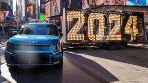 An image of a blue Kia EV9 with the 2024 numerals on a trailer behind it in Times Square, New York.