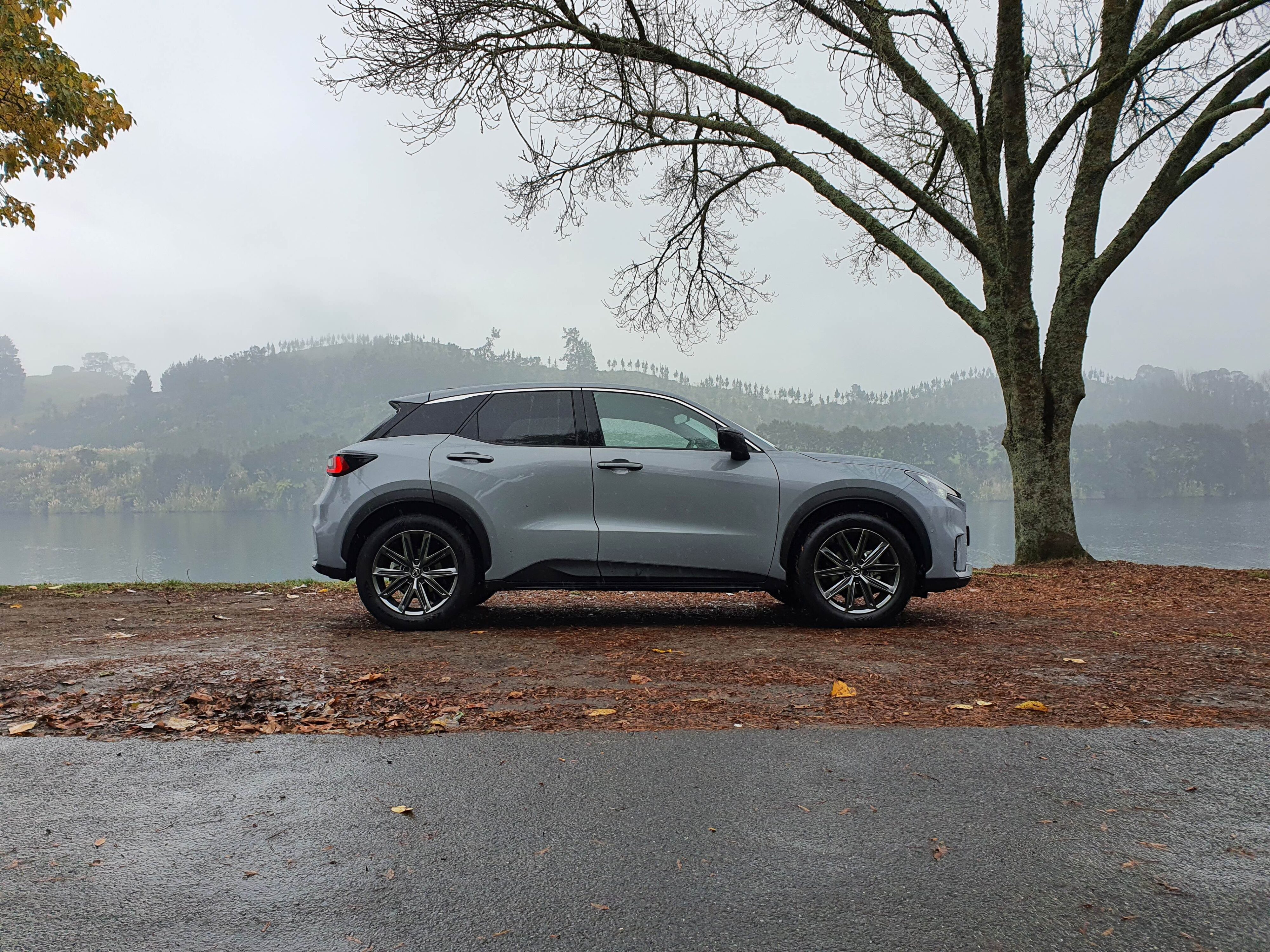 Side view of a 2024 Lexus LBX Relax AWD in Concrete Grey with bare trees, misty skies and Lake Karapiro in the background.