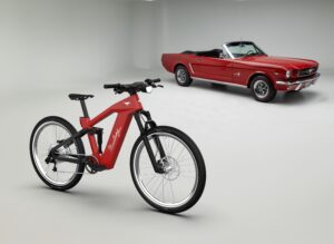 A promotional photo of a red Ford N+ Mustang inspired E-bike with a classic Ford Mustang convertible in the background.