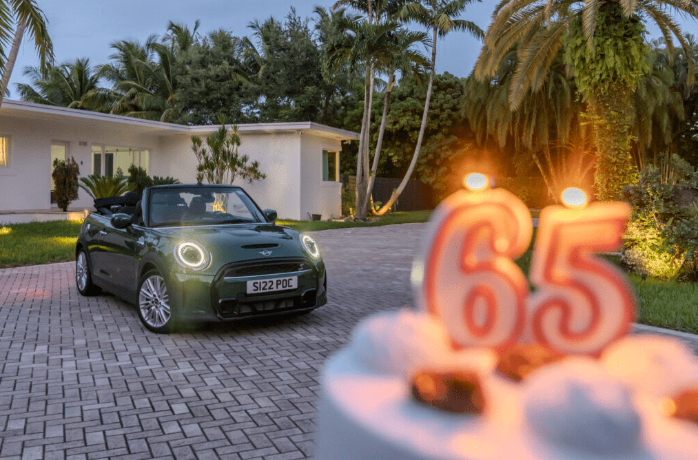 A Mini Cooper Cabriolet in the background and a birthday cake with 65 candles on it. Celebrating 65 years of Mini.