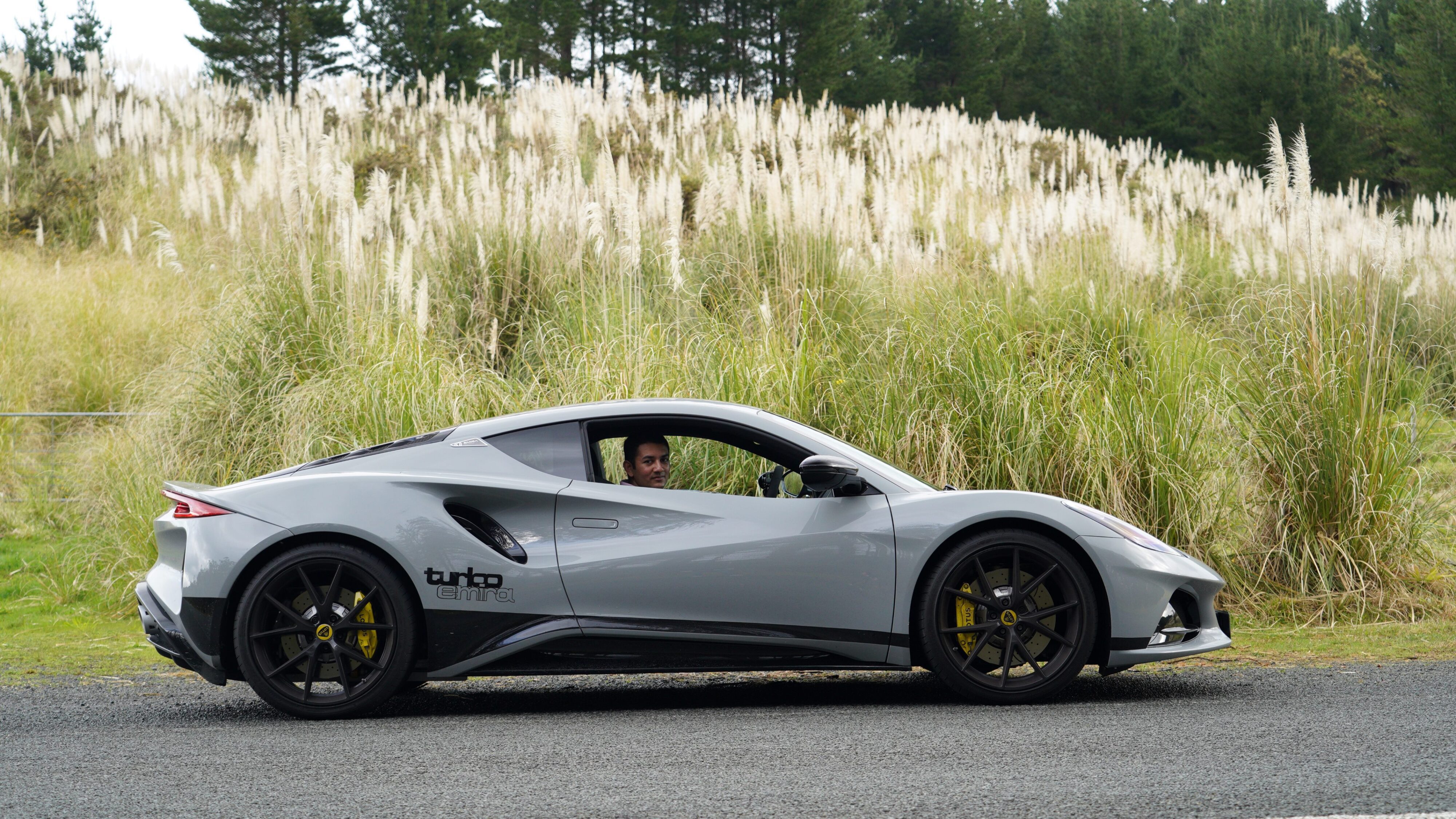 Side view of a 2024 Lotus Emira i4 Turbo in Nimbus Grey with Matthew D'Souza in the driver's seat.