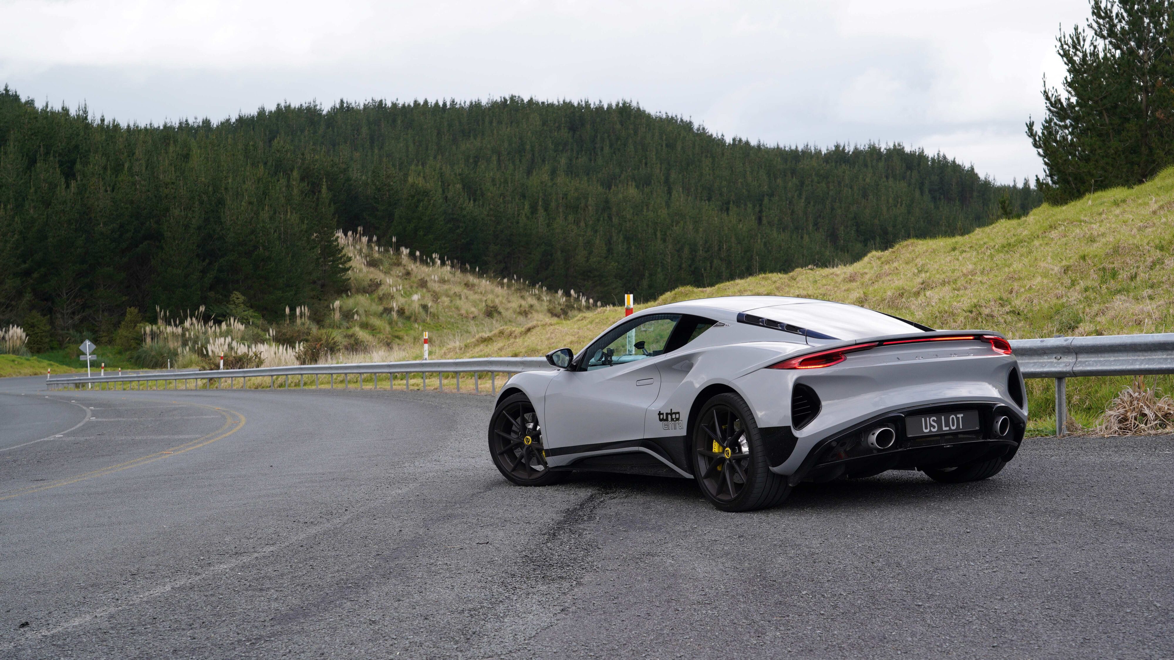 Rear three quarters view of a 2024 Lotus Emira i4 Turbo in Nimbus Grey with an s-bend and forest in the background.