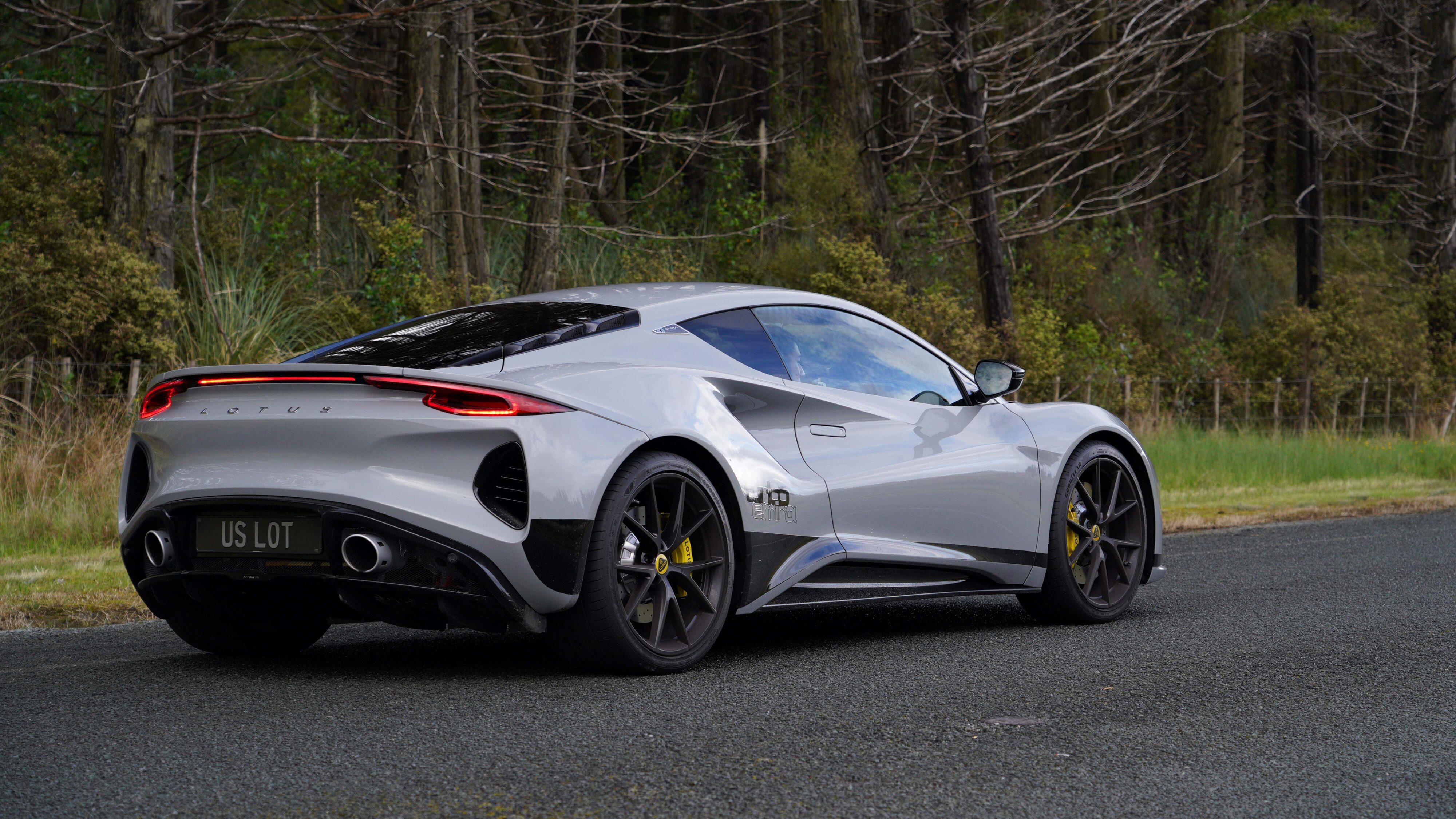 Rear three quarters view of a 2024 Lotus Emira i4 Turbo in Nimbus Grey with a forest in the background.