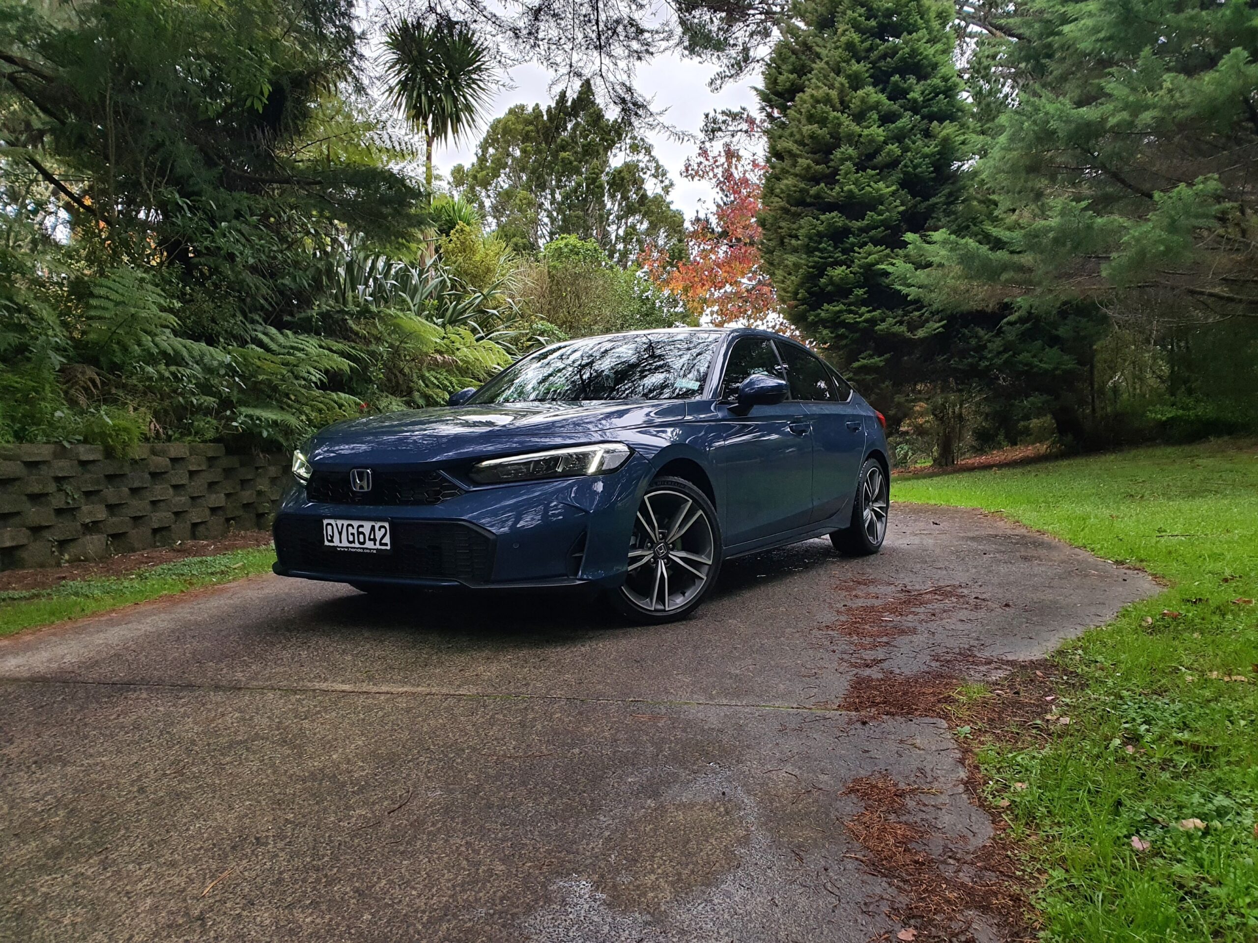 Front three quarters view of a 2025 Honda Civic e:hev in Ocean Blue Pearl with greenery in the background.