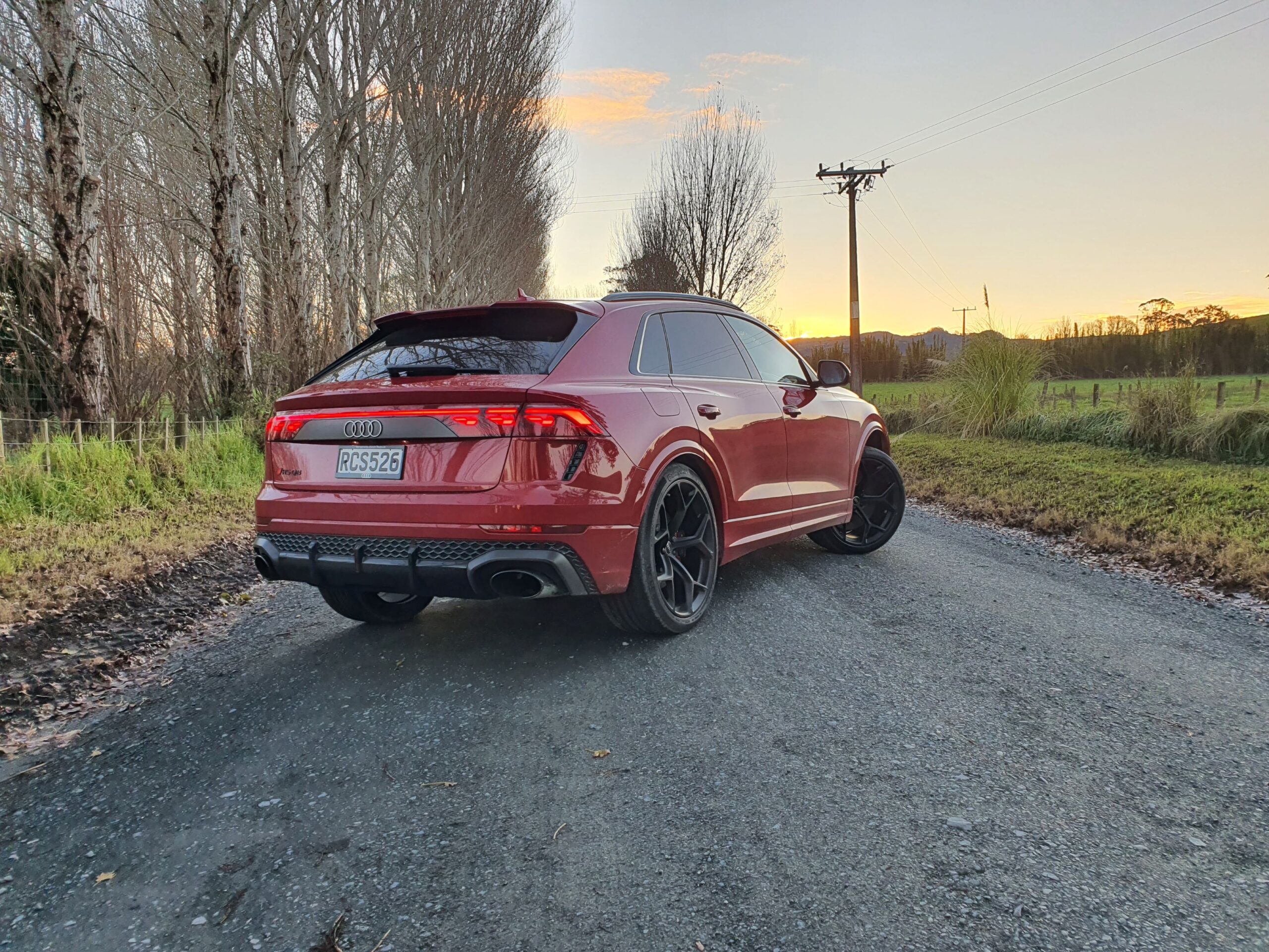 Rear three quarters view of a 2025 Audi RS Q8 Performance in Chili Red with the sun set and bare trees in the background.