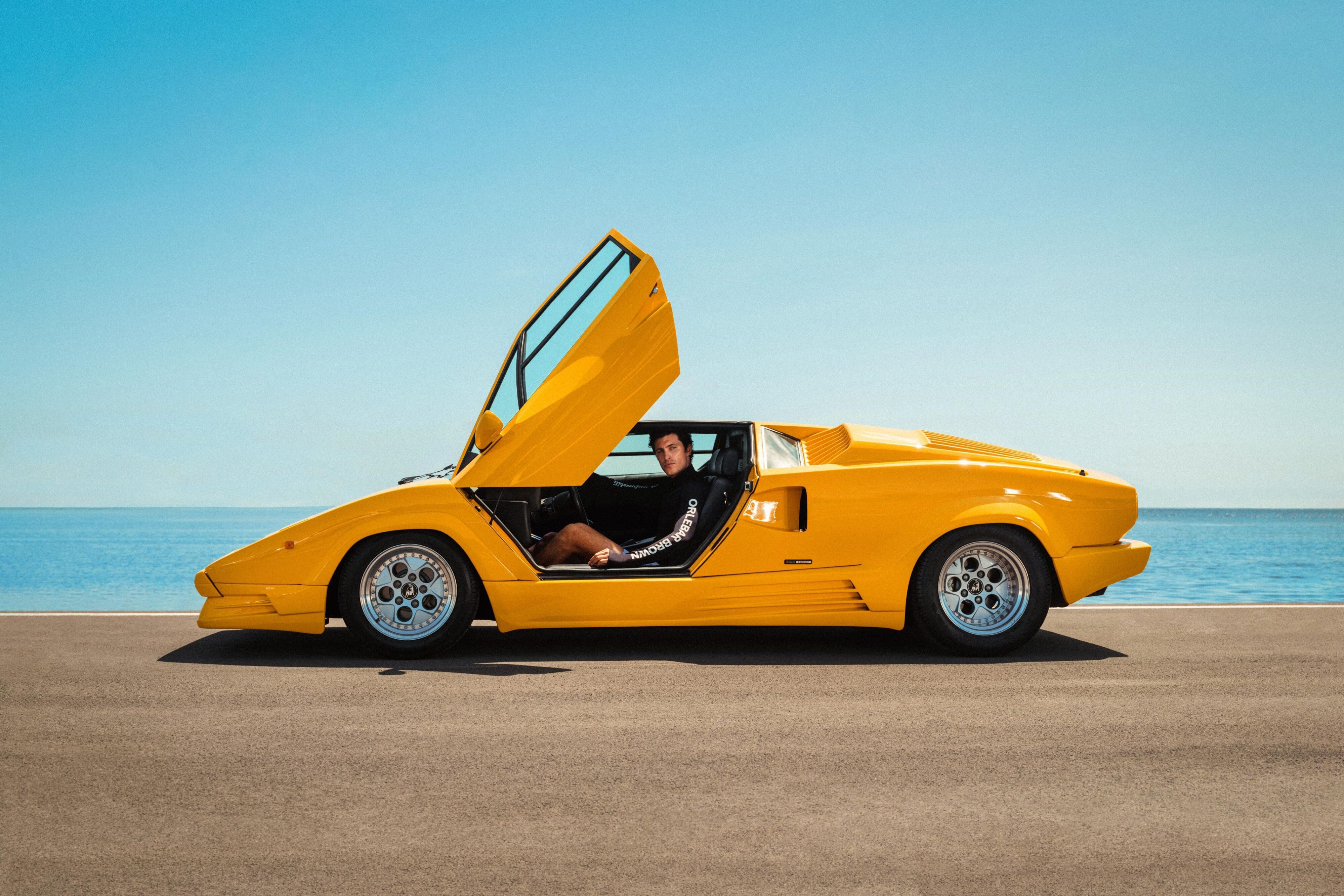 A photo of a model wearing the Orlebar Brown x Lamborghini collection while sitting in a classic yellow Lamborghini Countach.